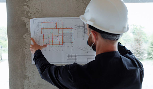 Construction worker in a white hard hat points to architectural blueprints taped to a concrete wall.