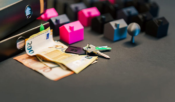 Euro banknotes and a set of house-shaped keycaps sit on a desk beside a few real keys and a keyboard, suggesting paying rent or buying a home.