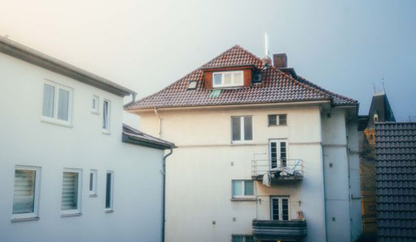 White multi-story residential building with a red tile roof and small balcony, flanked by neighboring houses.