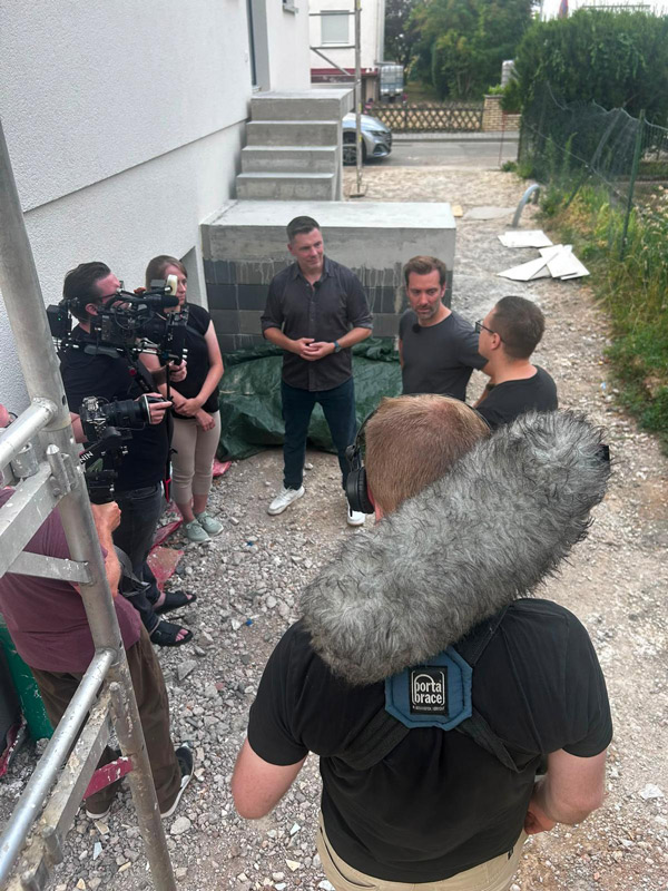 Film crew conducts an outdoor interview at a construction site, with a man speaking while others hold cameras and a boom mic nearby.